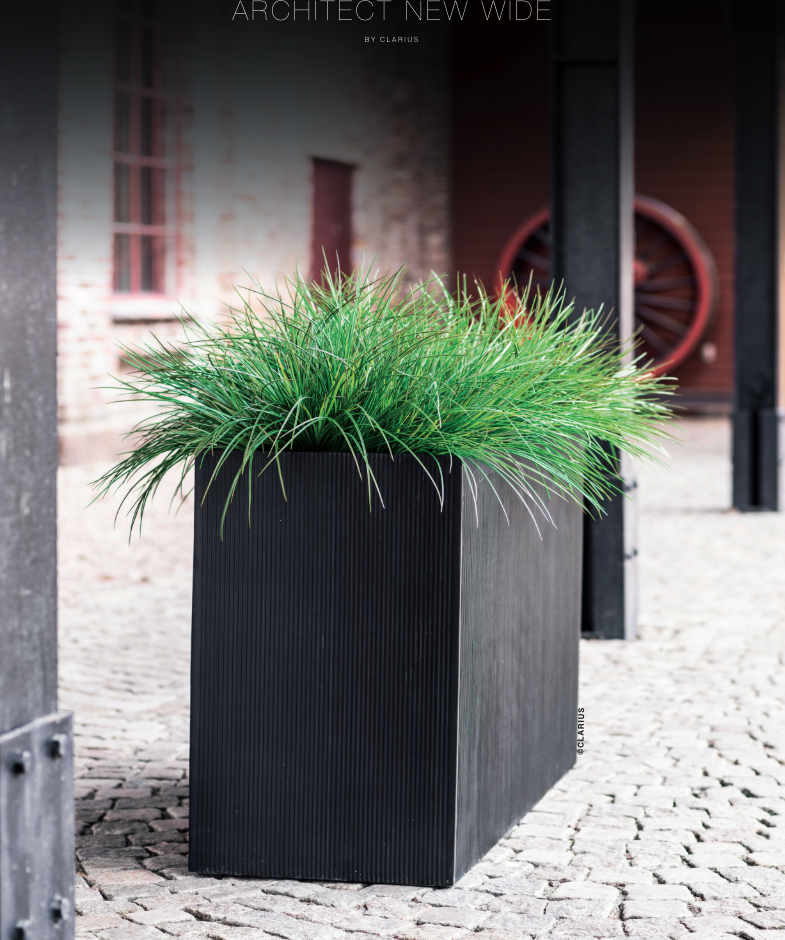 Svart rektangulär kruka med grönt gräs på utomhusplats med stenläggning och arkitektur i bakgrunden