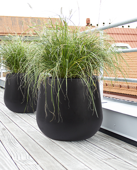 Svart kruka med hjul på terrass/balkong med gräs. Takrennor och byggnader synliga i bakgrund.